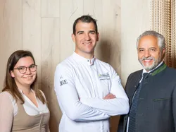Three smiling people including a chef in white uniform posing in front of wooden wall