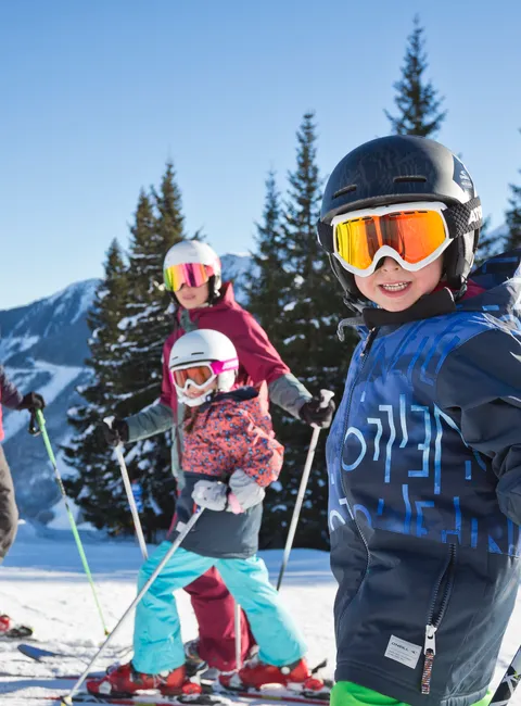 Family skiing on snowy mountain slope with ski lift and pine trees under blue sky