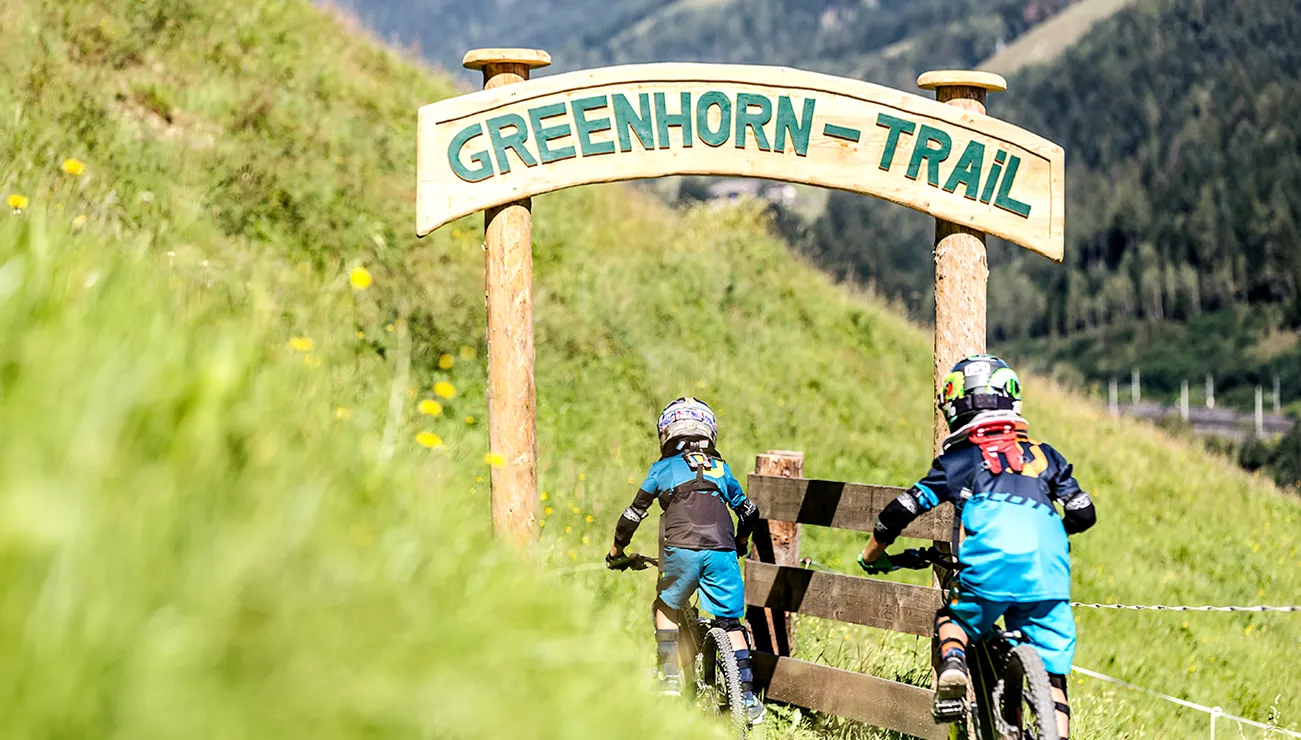 Zwei Kinder auf Mountainbikes fahren in den Greenhorn-Trail in alpiner Wiesenlandschaft