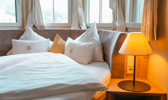 Cozy hotel bed with soft pillows and warm bedside lamp near corner windows