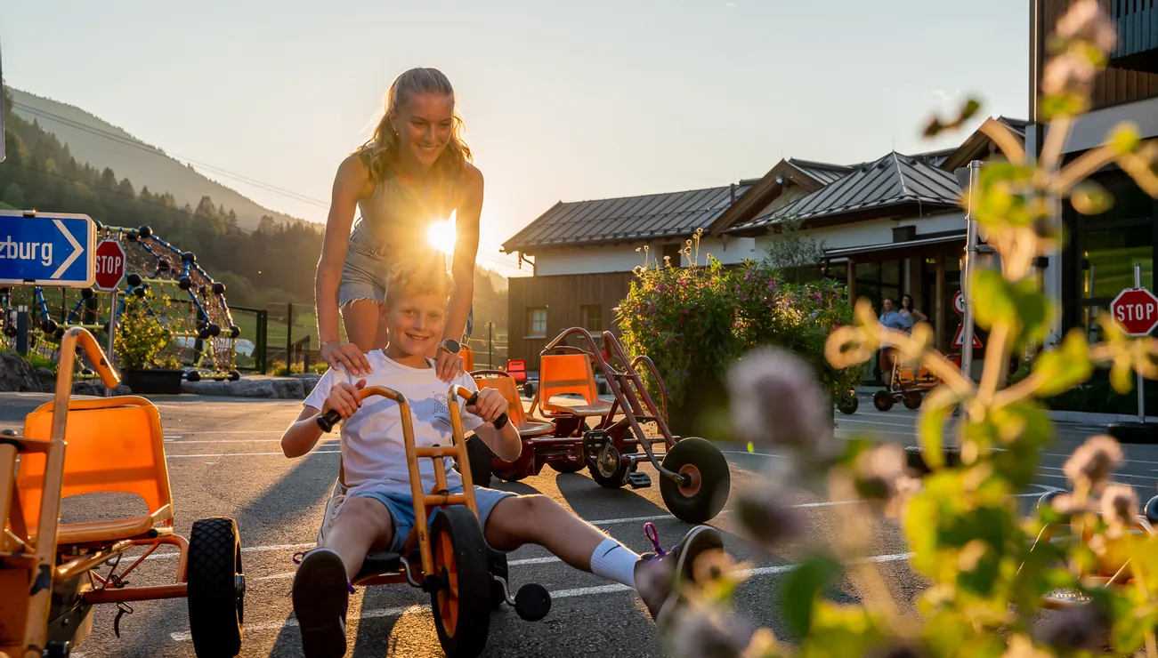 Junge fährt Gokart auf einem Parkplatz im Sonnenschein, begleitet von einem Mädchen
