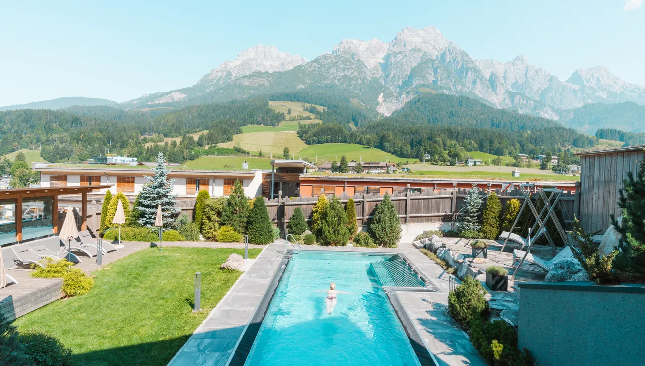 Außenpool mit Liegestühlen vor Alpenkulisse in sonniger Hotelanlage