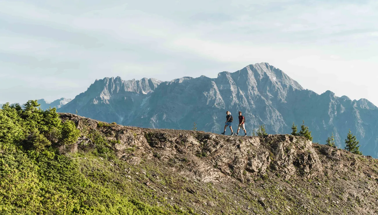 Zwei Wanderer gehen entlang eines Bergrückens mit alpinen Gipfeln im Hintergrund