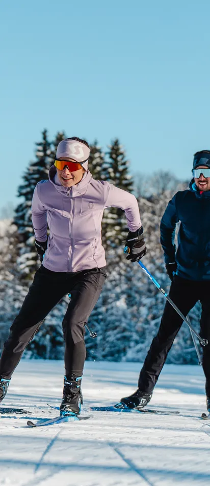 Zwei Personen beim Langlaufen auf verschneiter Loipe mit Alpen im Hintergrund