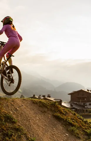 Frau in pinkem Outfit springt mit Mountainbike über Erdschanze bei Sonnenuntergang
