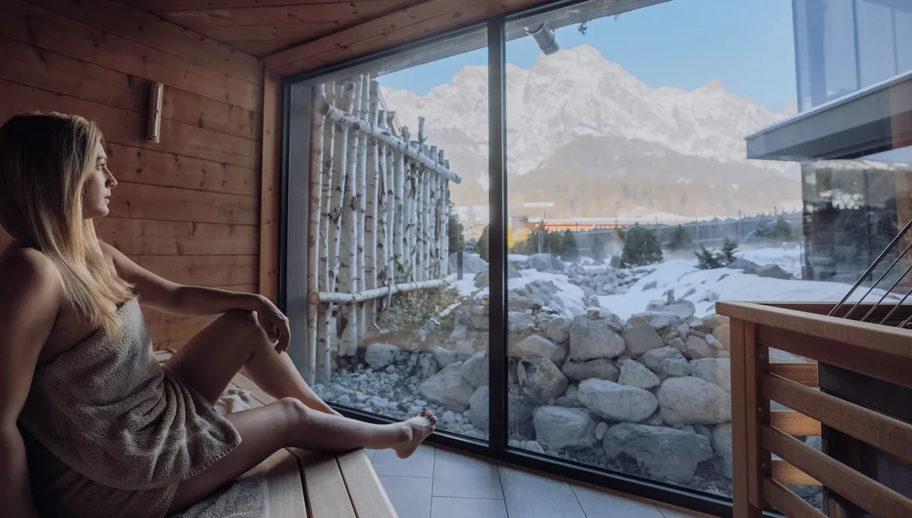 Frau in Sauna mit Blick auf verschneite Berglandschaft durch großes Fenster