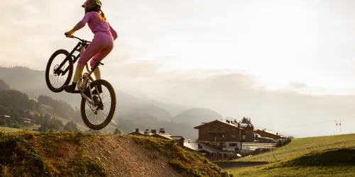 Frau in pinkem Outfit springt mit Mountainbike von Erdhügel im alpinen Sonnenaufgang
