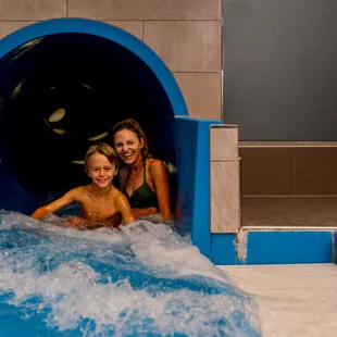 Smiling mother and son exiting indoor water slide into pool with blue splash