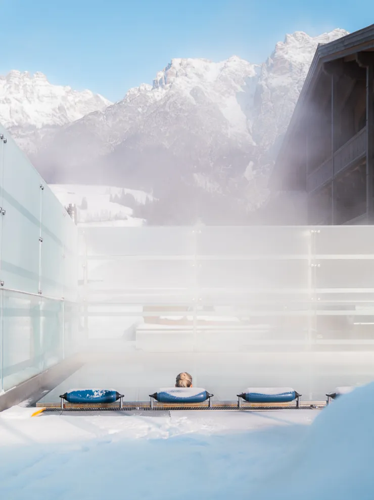 Person relaxing in outdoor spa pool with steam rising, snowy mountains in the background