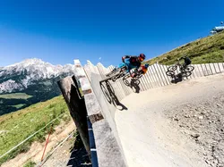 Mountainbiker fahren auf steiler Holzkurve im alpinen Bikepark mit Bergpanorama