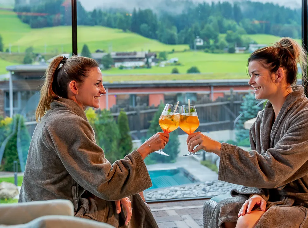 Two women in spa robes toasting drinks with mountain view in wellness resort