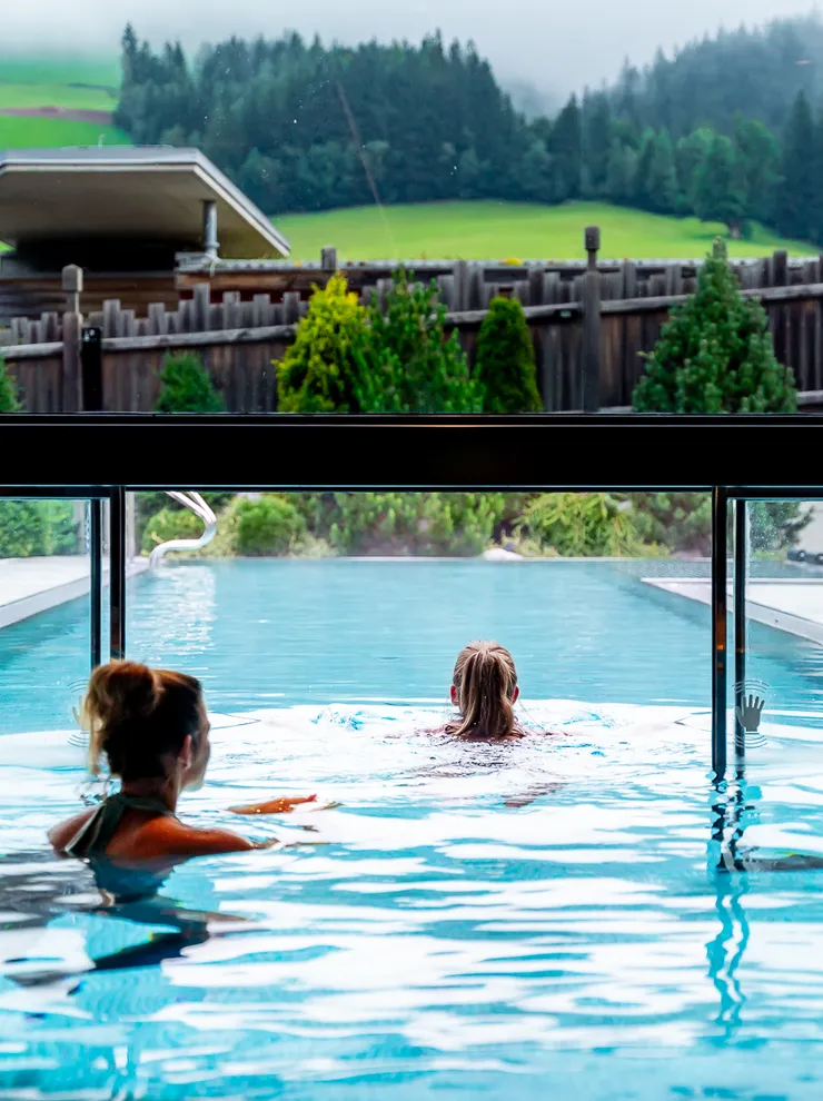 Zwei Frauen schwimmen in einem Infinity-Pool mit Blick auf grüne Hügellandschaft