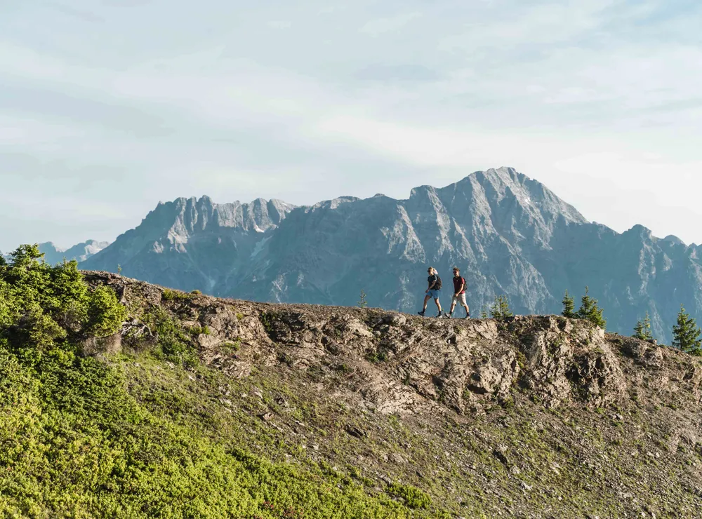 Zwei Wanderer gehen entlang eines Bergrückens mit alpinen Gipfeln im Hintergrund