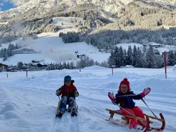 Zwei Kinder rodeln auf verschneitem Weg vor alpiner Bergkulisse