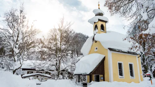 Snow-covered yellow chapel surrounded by winter trees and mountains in a serene village setting
