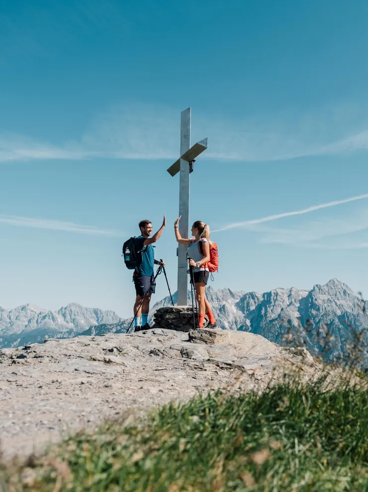 Wanderer geben sich High-Five am Gipfelkreuz mit Alpenpanorama im Hintergrund