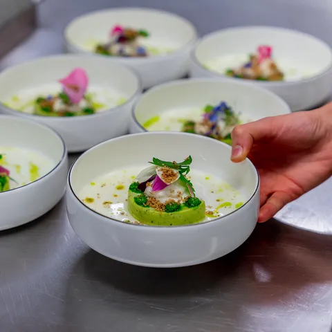 Gourmet appetizers in white bowls with vegetable mousse, herbs, and edible flowers on stainless steel counter