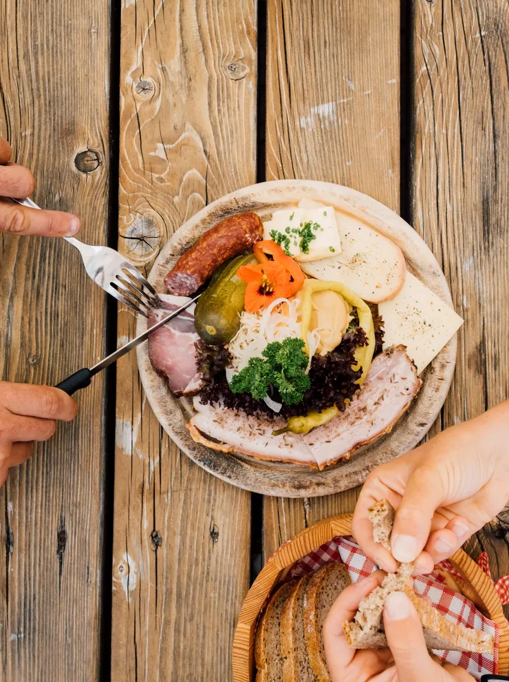 Rustikaler Holztisch mit traditioneller Fleisch-Käse-Platte und Händen, die Brot brechen