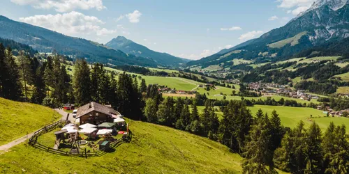 Berghütte mit Außensitzplätzen auf grüner Hügellandschaft mit Blick ins Alpental
