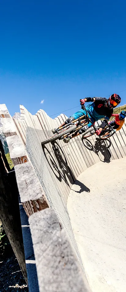 Mountain bikers riding on a steep wooden berm in an alpine bike park with mountain views