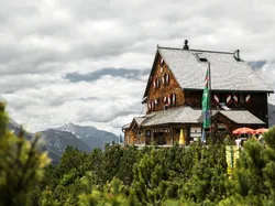 Alpenhütte mit Holzfassade und Fahnen, umgeben von Kiefern und wolkigem Himmel