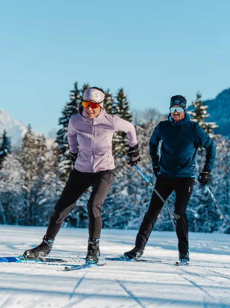 Zwei Personen beim Langlaufen auf verschneiter Loipe mit Alpen im Hintergrund