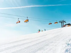 Skifahrer auf verschneiter Piste unter orangefarbenen Skiliften bei blauem Himmel