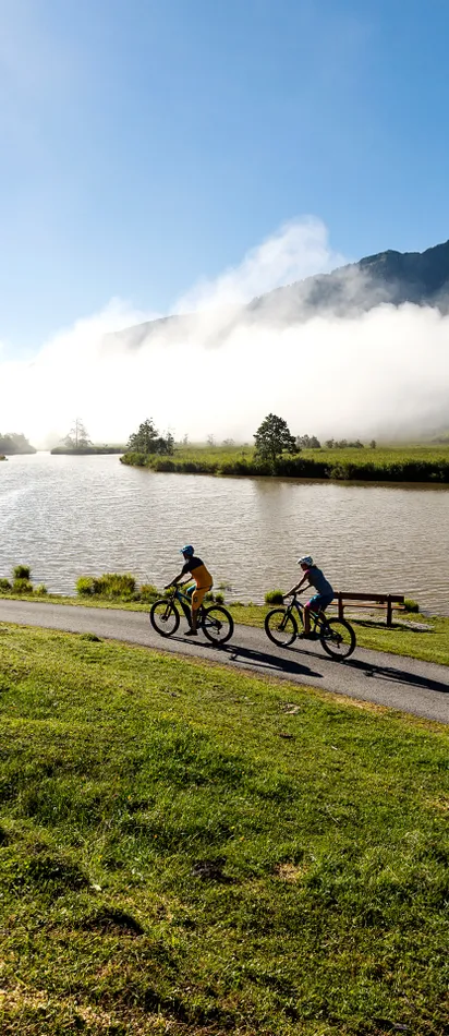 Radfahrer fahren an einem Bergsee entlang bei sonnigem Morgen mit Nebel