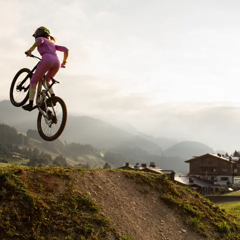 Mountain biker in pink gear doing a jump on dirt trail at sunset near alpine village