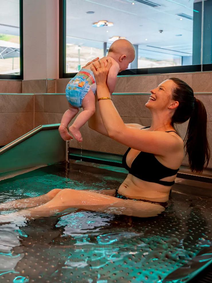 Lächelnde Frau im Bikini hält Baby im Innenpool eines Wellness-Spas hoch