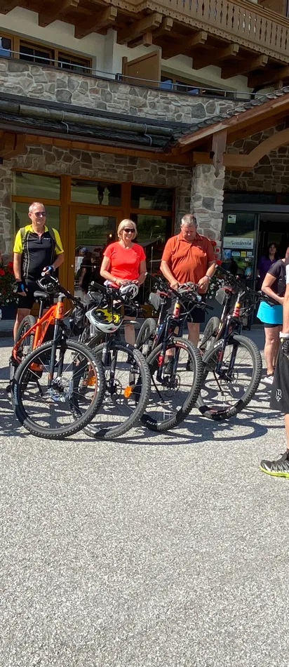 Group of cyclists with mountain bikes gathered outside a rustic lodge in sunny weather