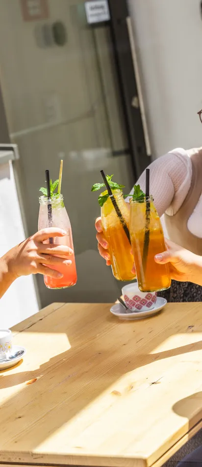 Drei Frauen in Tracht stoßen mit bunten Sommergetränken an einem sonnigen Tisch im Freien an