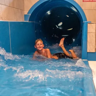 Smiling boy exits a blue water slide into a pool at an indoor water park