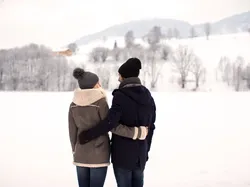 Paar in Winterkleidung blickt sich an vor verschneiter Landschaft mit Bergen