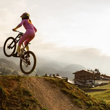 Frau in pinkem Outfit springt mit Mountainbike über Erdschanze bei Sonnenuntergang