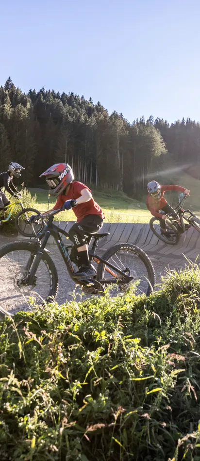 Kinder beim Mountainbiken auf einem Schotterweg mit Steilkurven in sonniger Waldlandschaft