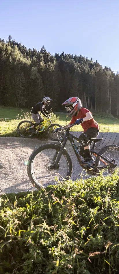 Kids mountain biking on a dirt trail with berms in a sunny forest landscape