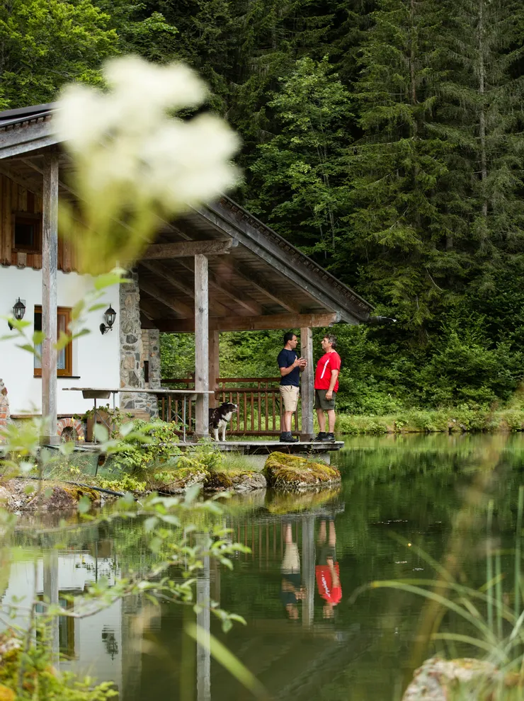Zwei Männer unterhalten sich auf der Veranda einer Holzhütte am Waldsee, Hund steht daneben