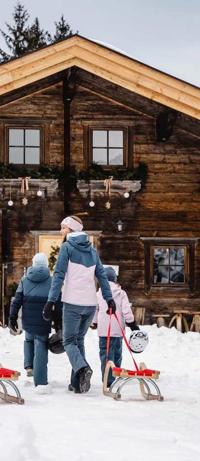 Family pulling sleds through snow toward rustic wooden mountain cabin