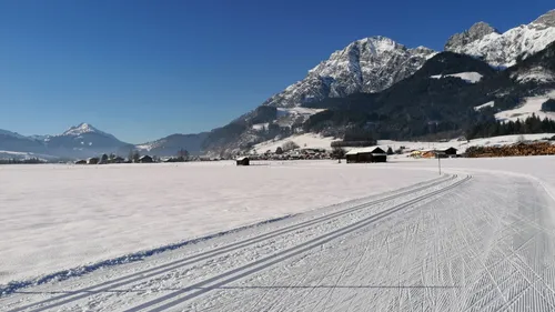 Langlaufloipe durch verschneite Landschaft mit Alpen und blauen Himmel im Hintergrund