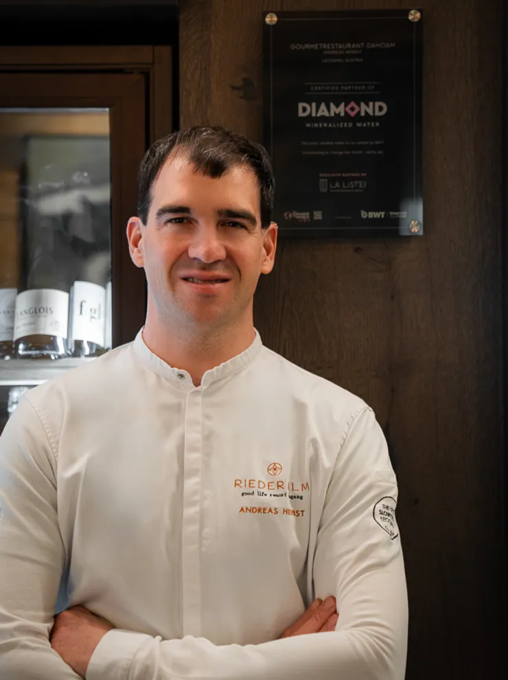 Male chef in white Riederalm uniform standing in front of dark wood panel with arms crossed