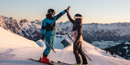 Zwei Skifahrer geben sich ein High Five auf einer verschneiten Piste bei Sonnenaufgang