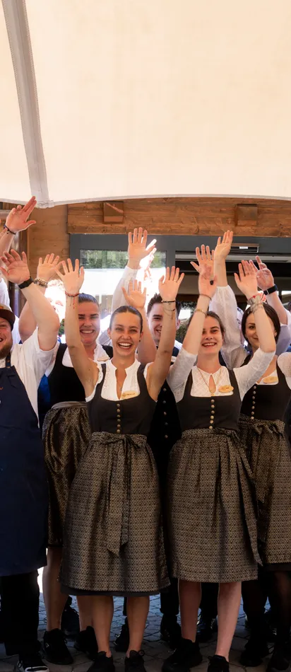 Lächelndes Restaurantteam in traditioneller bayerischer Tracht hebt draußen die Hände