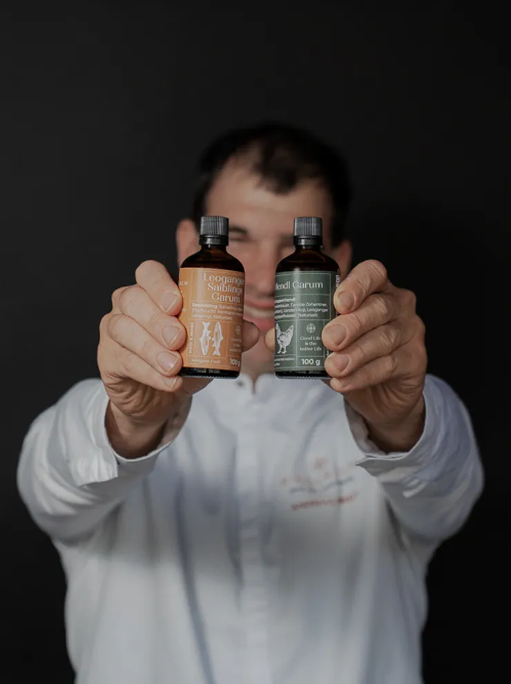 Man in white shirt holding two bottles of fermented Garum sauces against dark background