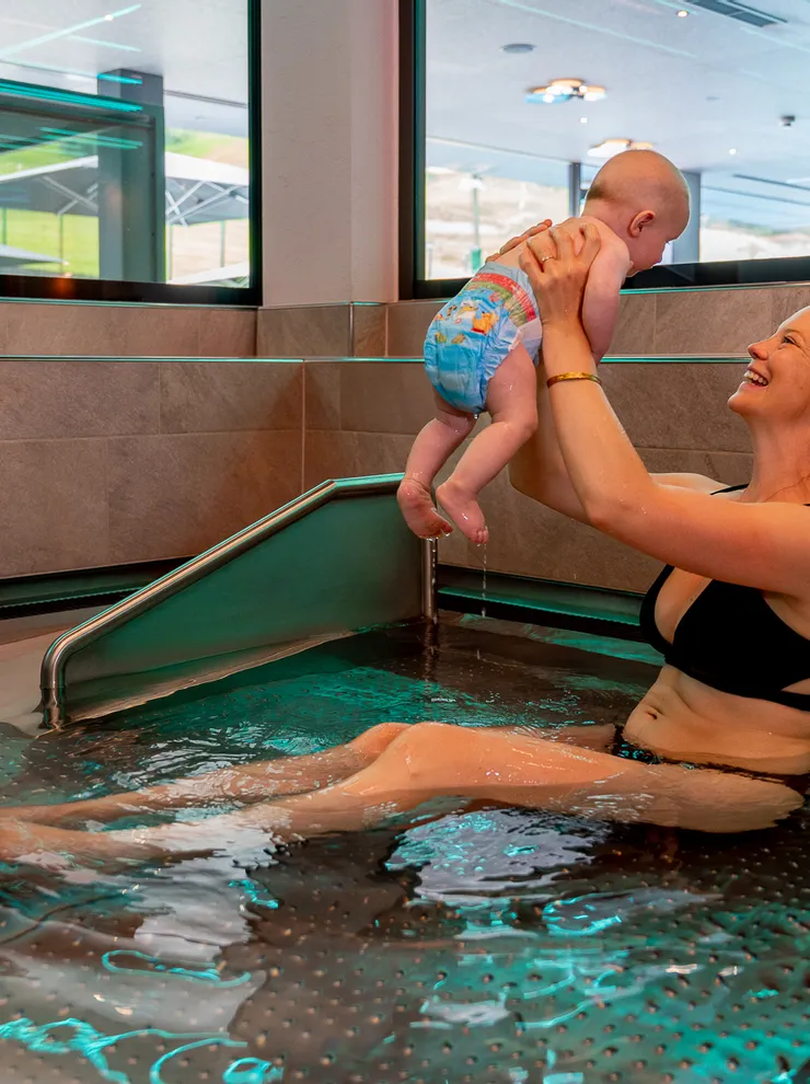 Lächelnde Mutter im Bikini hebt Baby in Schwimmwindel in einem Innenpool