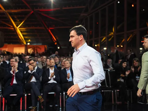 Mann in weißem Hemd betritt eine Bühne bei einer Business-Veranstaltung mit applaudierendem Publikum