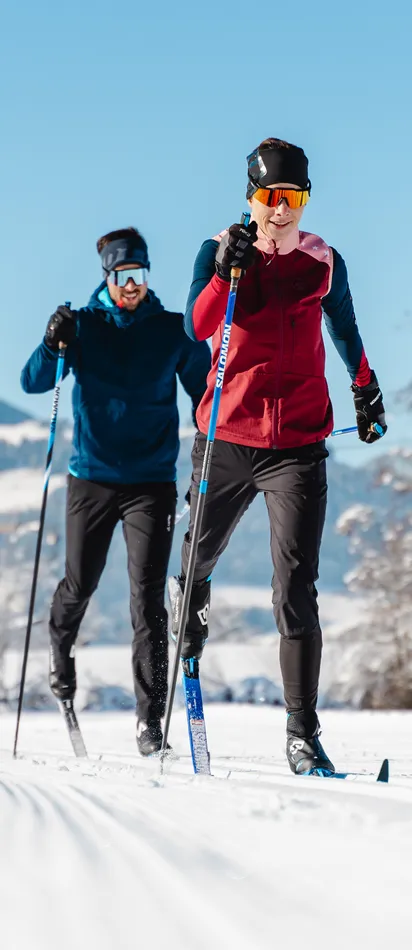 Zwei Langläufer auf einer verschneiten Loipe vor winterlicher Bergkulisse