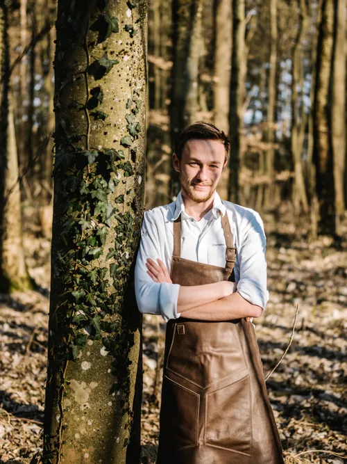 Junger Mann mit Lederschürze lehnt an efeubewachsenem Baum im sonnigen Wald