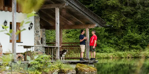Zwei Männer unterhalten sich auf der Veranda einer Hütte am Waldsee mit einem Hund in der Nähe