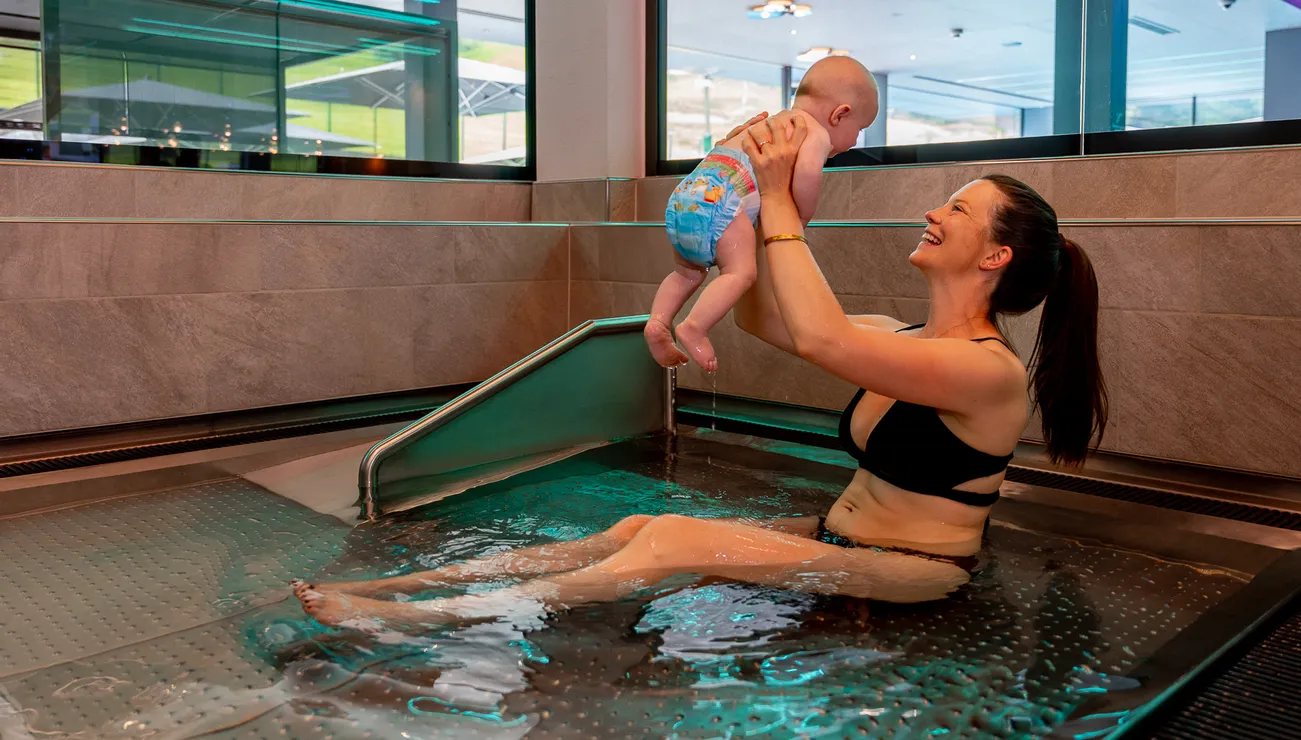 Lächelnde Mutter im Bikini hebt Baby in Schwimmwindel in einem Innenpool